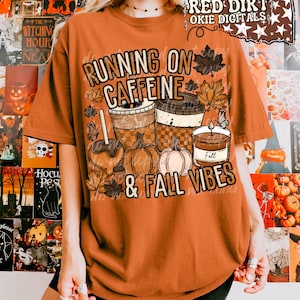 May include: A burnt orange t-shirt featuring a graphic print with the text RUNNING ON CAFFEINE & FALL VIBES. The design includes illustrations of two coffee cups, a caramel apple, two pumpkins, a candle labeled Fall, and autumn leaves.