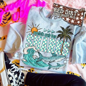May include: A light blue sweatshirt with a graphic that says "Watch Out For Sharks". The graphic includes a sun, palm tree, ocean wave, and sharks. The sweatshirt is on a pink floral patterned surface.