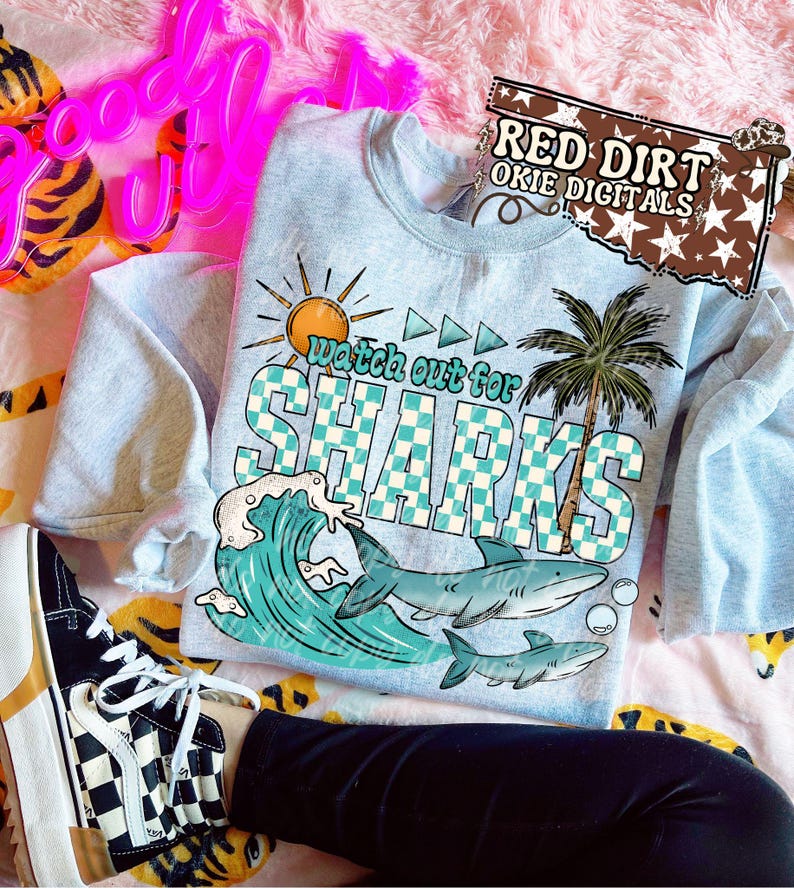 May include: A light blue sweatshirt with a graphic that says "Watch Out For Sharks". The graphic includes a sun, palm tree, ocean wave, and sharks. The sweatshirt is on a pink floral patterned surface.