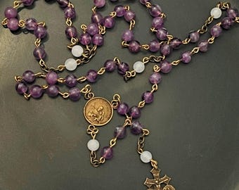 May include: A rosary with purple amethyst beads and gold-colored metal chain. The rosary features a crucifix pendant and a circular medal with religious figures. The beads are interspersed with white beads.