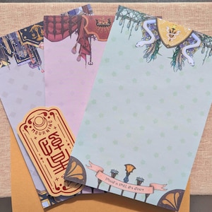 May include: Three decorative paper notepads with different designs. The top notepad has a green background with a white snake design and the text "Don't tell a soul". The middle notepad has a pink background with a red, white, and blue design. The bottom notepad has a white background with a red and black design and the text "Mourning Dove".