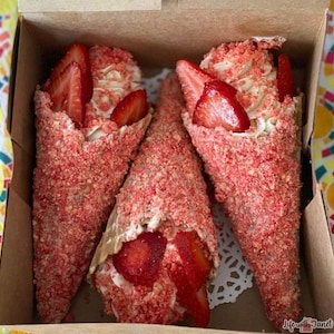 May include: Three waffle cones filled with whipped cream and strawberries, covered in red and white sprinkles. The cones are in a cardboard box.