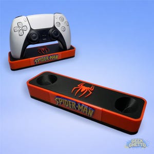 Può includere: Un controller di gioco bianco e nero poggia su una base rossa e nera con le parole "SPIDER-MAN" e un logo a forma di ragno. È visibile anche una seconda base con due ritagli circolari.