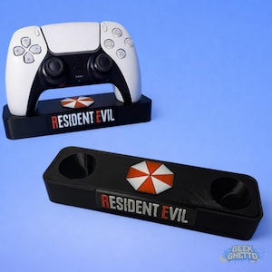 Puede incluir: Un mando de juego blanco y negro descansa sobre un soporte negro con el texto "RESIDENT EVIL" en rojo y blanco. El soporte también presenta un logotipo de paraguas. También hay otro soporte con el mismo diseño.