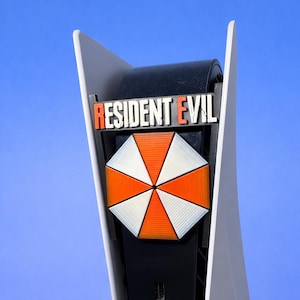 Può includere: Una console di gioco in bianco e nero con le parole "RESIDENT EVIL" in lettere rosse e bianche. Sotto, un design geometrico di un ombrello arancione e bianco.