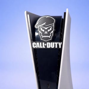 Peut inclure: Console de jeu blanche et noire avec le logo Call of Duty. Le logo représente un crâne portant un béret. Le texte "CALL OF DUTY" est en dessous du crâne. La console a un design élégant et moderne.