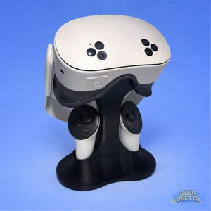 Op de afbeelding: Een witte VR-headset met zwarte accenten en een bijpassende zwarte standaard. De standaard houdt de headset en twee witte controllers met zwarte knoppen vast. De headset heeft meerdere kleine zwarte cameralenzen aan de voorkant.
