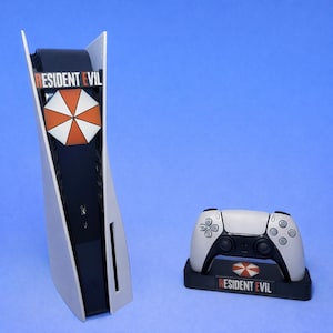 May include: A white and black gaming console with a matching controller and stand. The console and controller feature the text "RESIDENT EVIL" and a red, white, and orange umbrella logo. The stand also has the text "RESIDENT EVIL".