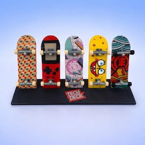 Peut inclure: Ensemble de cinq mini-skateboards aux motifs variés, dont une manette de jeu rouge et noire, un personnage de dessin animé jaune et un skateboard avec le texte "Tech Deck". Les planches sont présentées sur un présentoir noir.
