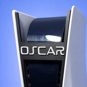 Può includere: Primo piano di una console di gioco bianca e nera con il nome "OSCAR" in lettere bianche. La console ha una sezione centrale curva blu scuro e un design elegante e moderno. Lo sfondo è una sfumatura di blu.