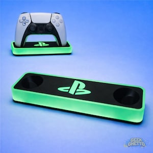 以下が含まれることがあります： 白と黒のPlayStation 5コントローラーが、PlayStationのロゴが入った緑色に光るスタンドの上に置かれています。下には、同じロゴと緑色の照明が付いた黒い充電ステーションが見えます。背景は青のグラデーションです。