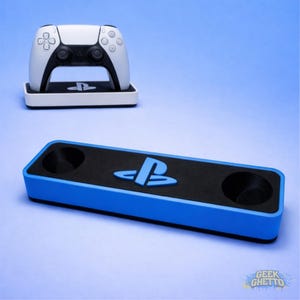 May include: A white and black PlayStation 5 controller sits on a white base with the PlayStation logo. Below, a blue and black charging station features the PlayStation logo and two charging ports. The background is a gradient blue.
