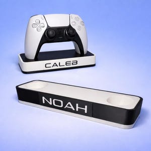 May include: A white and black gaming controller on a matching stand with the name "CALEB". Below is a white and black charging station with the name "NOAH". The items are on a blue background. The items are for a gaming console.