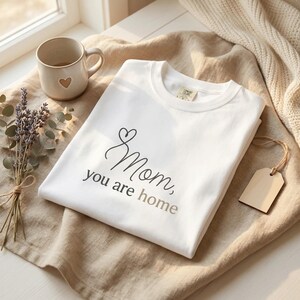 Puede incluir: Una camiseta blanca con las palabras "Mom, you are home" en fuente cursiva. La camiseta está doblada sobre una superficie beige, junto a una taza con un corazón y un ramo de lavanda.