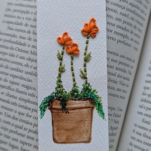 Könnte beinhalten: Bestickter Lesezeichen mit einer Aquarellillustration von drei orangefarbenen Blumen in einem braunen Topf mit grünen Blättern. Das Lesezeichen ist rechteckig und liegt auf einem offenen Buch.