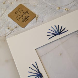 Pode incluir: Um passe-partout retangular branco com padrões em forma de leque bordados em azul escuro ao longo das bordas. Uma pequena etiqueta quadrada castanha com o texto DT's Embroidery Atelier é visível perto do canto superior esquerdo.