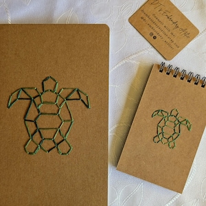 Pode incluir: Um caderno castanho e um bloco de notas castanho mais pequeno com espiral, cada um com um design geométrico de tartaruga marinha bordado a verde. Está incluído um pequeno cartão de visita retangular com o texto DT's Embroidery Atelier.