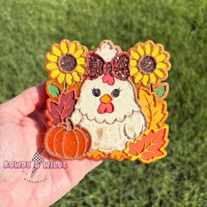 May include: A square decorative plaque featuring a white chicken with a glittery maroon bow, flanked by two yellow sunflowers. The scene includes an orange pumpkin and autumn leaves in shades of red, yellow, and orange. The text Rowdy & Wilde Mercantile is visible.