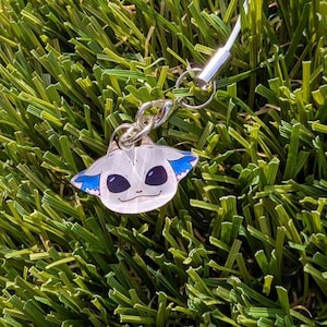 May include: A silver-toned keychain charm featuring a cartoon-style character with large, dark eyes and blue ear accents. The charm is attached to a silver chain and clasp, set against a backdrop of green artificial grass.