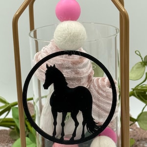 May include: A decorative glass vase with a black horse silhouette inside a circular frame. The vase is filled with pink fabric and white and pink felt balls. A gold metal frame surrounds the vase, topped with a pink sphere and a white felt ball.