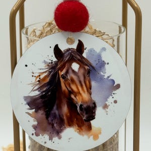 May include: A round ornament featuring a watercolor painting of a brown horse head with a white blaze. The ornament has a red felt ball attached to the top. The background has blue and purple watercolor splashes.