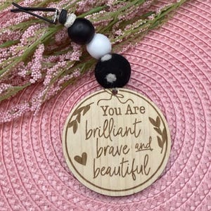 May include: A wooden car charm with the words "You Are brilliant brave and beautiful" engraved on it. The charm has a string of black, white, and gray beads. The background is a pink woven surface.