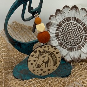 May include: A wooden horse pendant with a detailed horse head design and heart accents. The pendant is attached to a beaded strand with orange, cream, and brown beads. The pendant is displayed near a decorative sunflower and a teal metal sculpture.