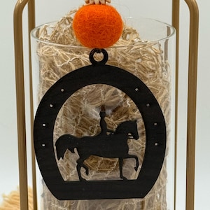 May include: A decorative glass vase filled with twine, featuring a black horseshoe-shaped cutout with a horse and rider silhouette. An orange felt ball sits atop the vase, suspended within a gold-colored metal frame. The overall theme is equestrian.