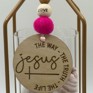 May include: A decorative hanging ornament featuring a wooden disc with the word "Jesus" and the phrases "The Way, The Truth, The Life." It includes a pink felt ball, a wooden bead with "LOVE" and a gold metal frame.