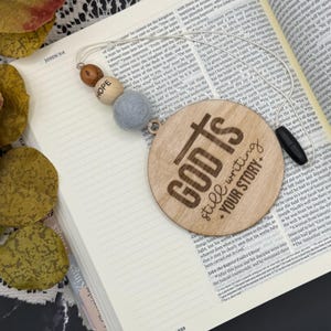 May include: Wooden bookmark with the text "GOD IS still writing YOUR STORY" in a circular design. The bookmark has a string with wooden beads and a gray felt ball. The bookmark is placed on an open book.