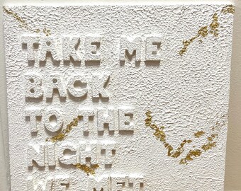 May include: White canvas wall art with the text "Take me back to the night we met" in raised gold letters. The background has a textured, rough surface with gold glitter accents.