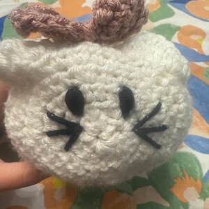 May include: A handmade, white crocheted bunny head with black eyes and whiskers. The bunny has a pair of pinkish-brown ears. The craft project is made with a textured yarn, giving it a soft, plush appearance. The bunny is a cute, whimsical item.