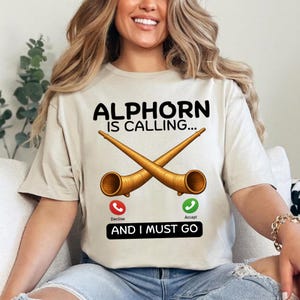 May include: Beige t-shirt with the text "ALPHORN IS CALLING..." above crossed alphorns. Below the horns are phone icons with "Decline" and "Accept" labels, and the phrase "AND I MUST GO".