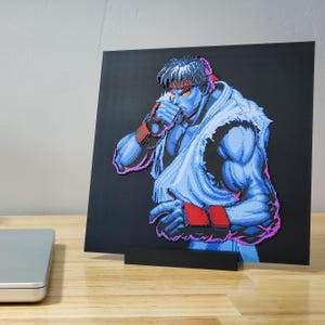 Könnte beinhalten: Ein Pixel-Art-Druck eines blauen und weißen Charakters mit einem roten Stirnband, einem weißen Gi und roten Handschuhen. Der Charakter befindet sich in einer Kampfpose mit einer Hand, die zum Gesicht erhoben ist.