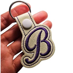 May include: Cream-coloured keyring featuring a purple embroidered letter "B". The letter is stitched onto a textured, off-white leather-like material. A silver metal ring and a white snap button secure the keyring.