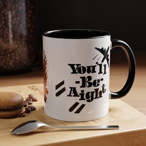May include: White ceramic mug with a black interior and handle. The mug features the text "You'll -Be- Aight" in black, along with a graphic of an airplane. A spoon and a macaron are on a wooden surface.