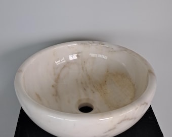 May include: A white marble vessel sink with subtle brown veining. The oval-shaped sink has a smooth, polished finish and a central drain hole. It is displayed on a black surface against a light gray background.