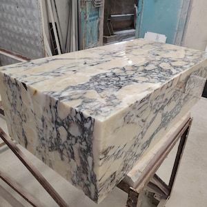 May include: A rectangular block of marble with a polished surface. The marble features a cream-colored base with intricate veining in shades of gray and black. The block is displayed on a metal frame.