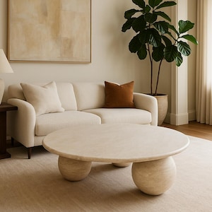 May include: A neutral-toned living room featuring a cream-colored sofa with two pillows and a brown accent pillow. A round, light-colored coffee table sits on a beige rug. A large framed abstract painting hangs on the wall.