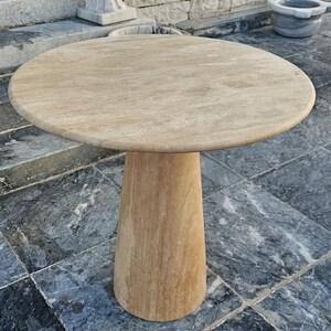 May include: A round, light-colored stone table with a single, conical base. The tabletop is approximately 36 inches in diameter. The table is made of a light beige stone and sits on a tiled surface.