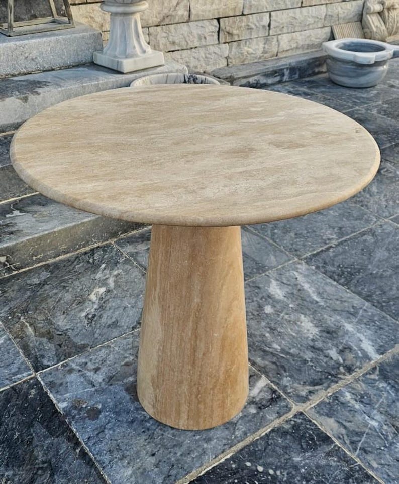 May include: A round, light-colored stone table with a single, conical base. The tabletop is approximately 36 inches in diameter. The table is made of a light beige stone and sits on a tiled surface.