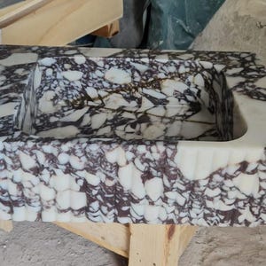 May include: Rectangular marble sink with a unique pattern of white and dark purple veining. The sink has a scalloped edge design and a curved interior. The sink is made of stone and is a bathroom fixture.