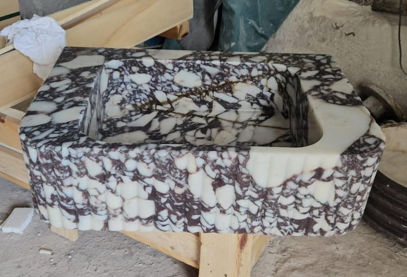 May include: Rectangular marble sink with a unique pattern of white and dark purple veining. The sink has a scalloped edge design and a curved interior. The sink is made of stone and is a bathroom fixture.