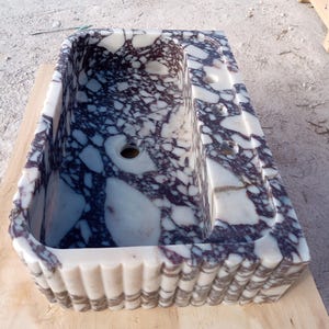Puede incluir: Un lavabo rectangular de m&aacute;rmol con un dise&ntilde;o de patr&oacute;n morado y blanco. El lavabo tiene una pila, un &aacute;rea elevada con tres agujeros y un borde decorativo festoneado. El lavabo es de piedra.