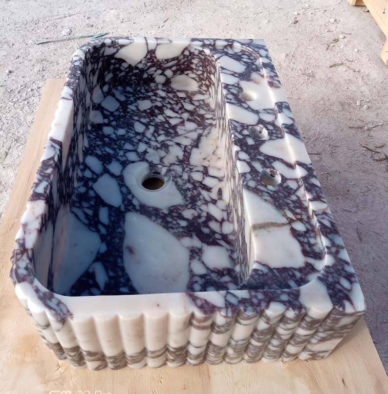 Puede incluir: Un lavabo rectangular de m&aacute;rmol con un dise&ntilde;o de patr&oacute;n morado y blanco. El lavabo tiene una pila, un &aacute;rea elevada con tres agujeros y un borde decorativo festoneado. El lavabo es de piedra.