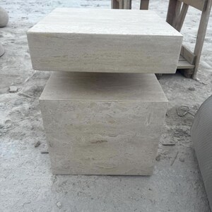 Peut inclure: Une table d'appoint en pierre beige clair avec un plateau rectangulaire et une base en forme de cube. La table est faite d'une pierre naturelle et a un design simple et moderne. La table mesure environ 61 cm de hauteur.