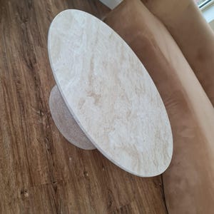 May include: Oval-shaped coffee table with a light beige marble-like top and a matching cylindrical base. The table is set on a dark brown wooden floor, with a brown sofa visible in the background. The table's design is modern and minimalist.