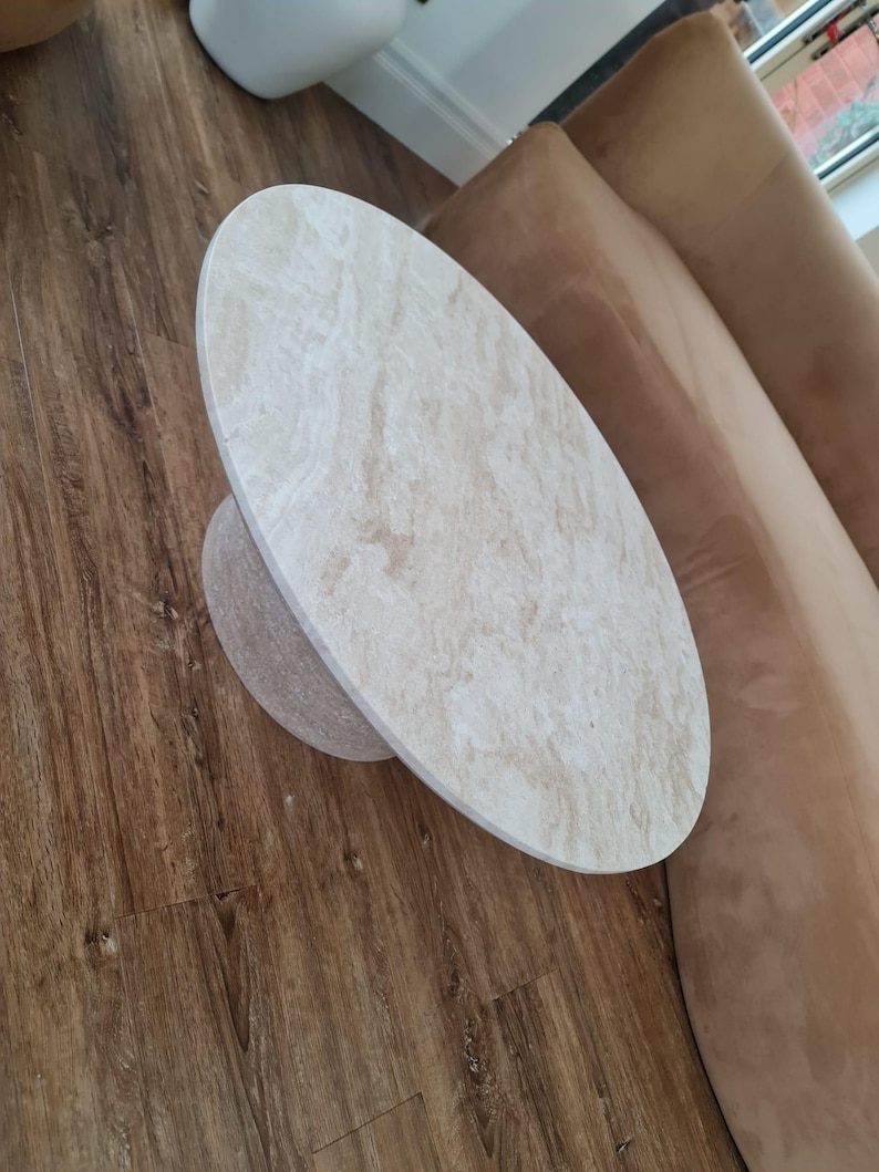 May include: Oval-shaped coffee table with a light beige marble-like top and a matching cylindrical base. The table is set on a dark brown wooden floor, with a brown sofa visible in the background. The table's design is modern and minimalist.