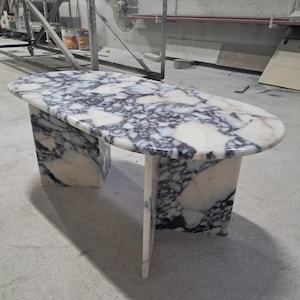 May include: Oval marble table with a white and dark grey pattern. The table has two rectangular supports and a smooth, polished surface. The design is modern and suitable for various interior settings.