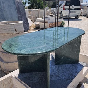 Puede incluir: Una mesa de centro de m&aacute;rmol verde con una tapa rectangular redondeada y dos patas de pedestal. La mesa est&aacute; hecha de m&aacute;rmol pulido con vetas naturales. La mesa est&aacute; al aire libre en un patio de piedra.